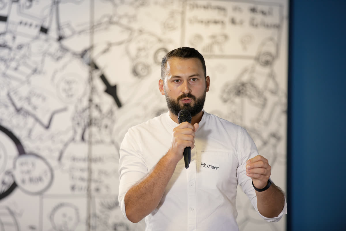 Cristian Pogan speaking at a demo day, microphone in hand, in front of an illustrated wall